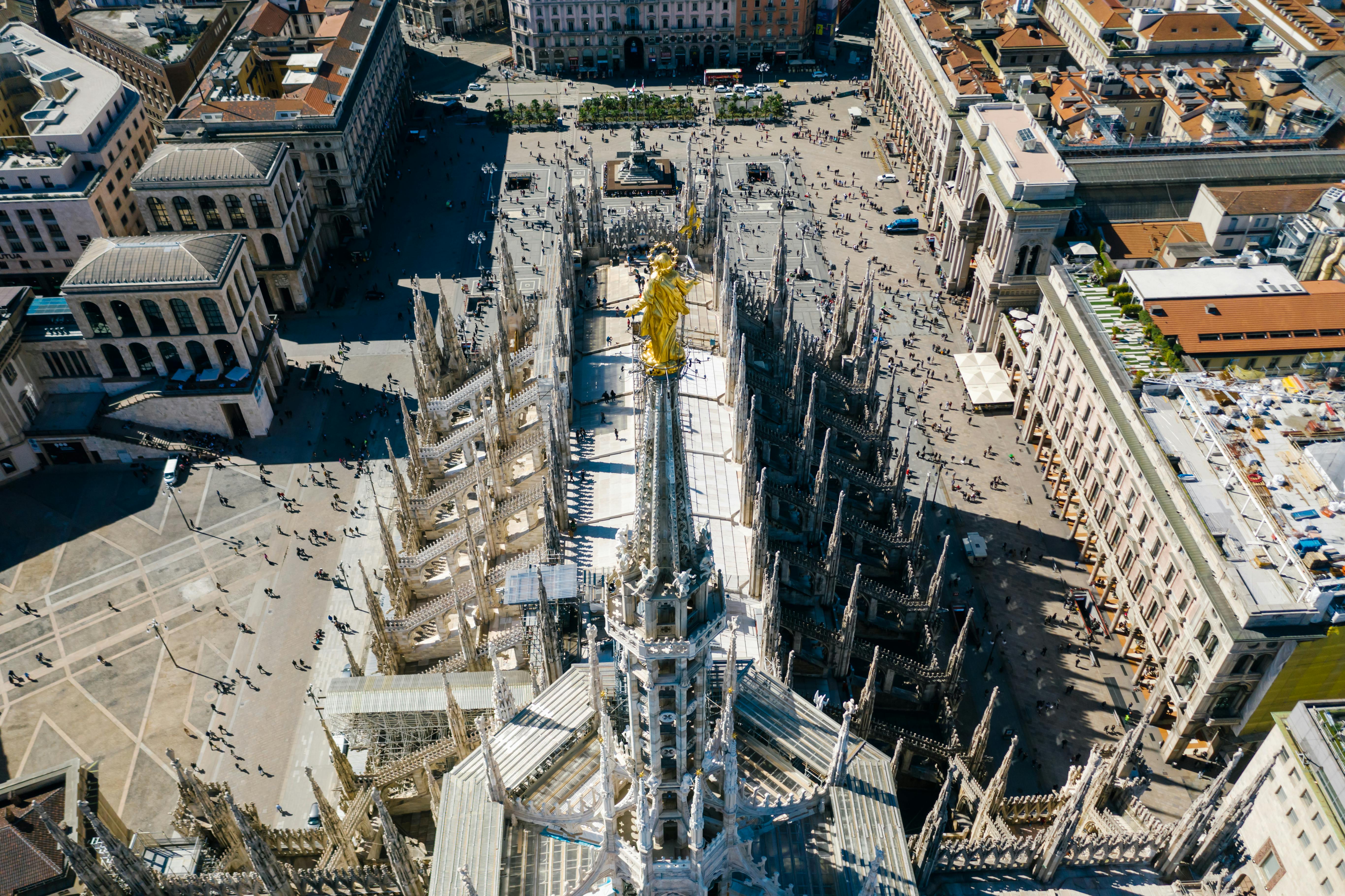 Aerial view of Milan Cathedral rooftop with detailed spires and golden statue overlooking the busy Piazza del Duomo.