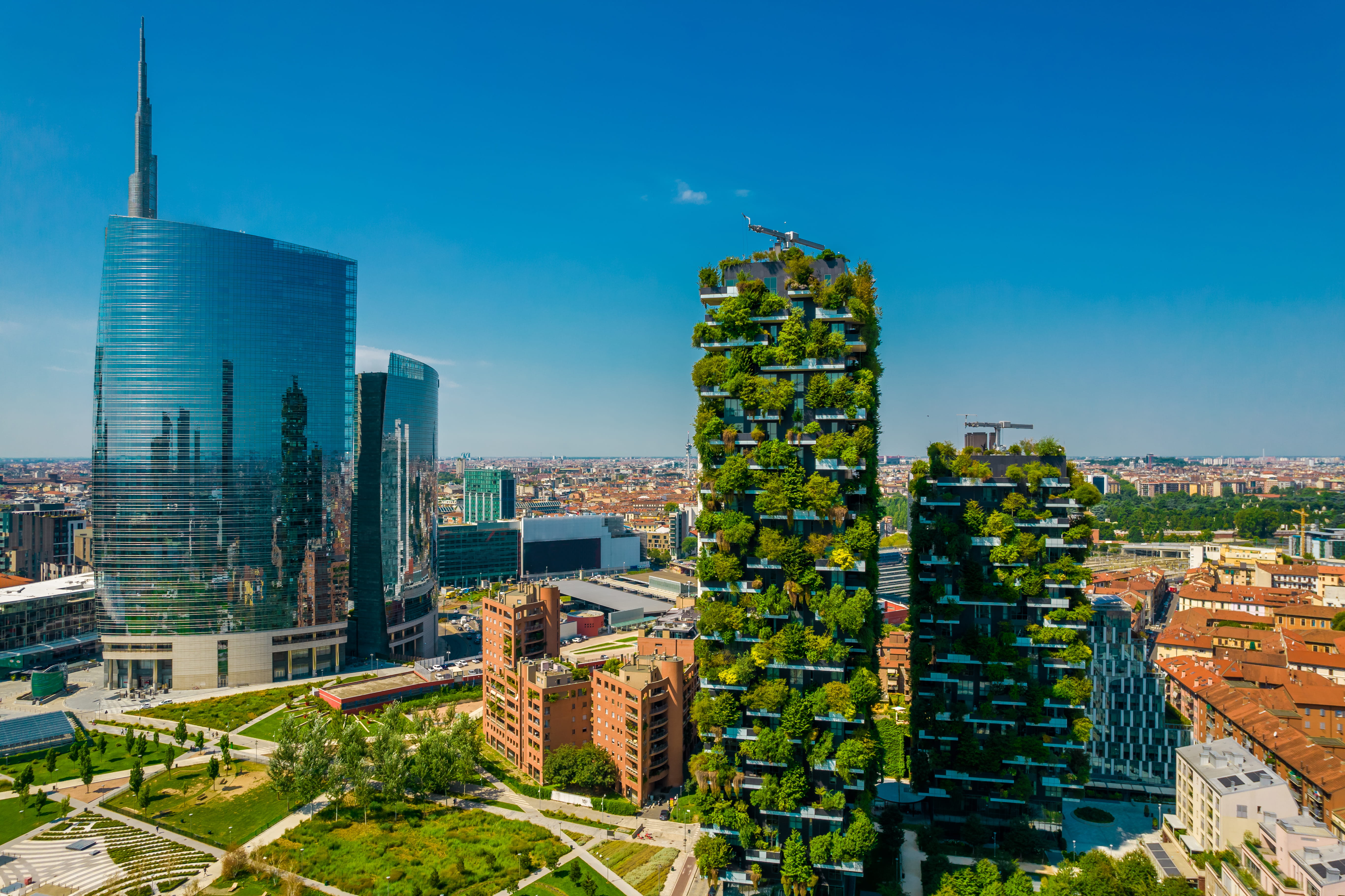 Vertical forest skyscrapers covered in dense greenery beside modern glass high-rise buildings in an urban cityscape under a clear blue sky.