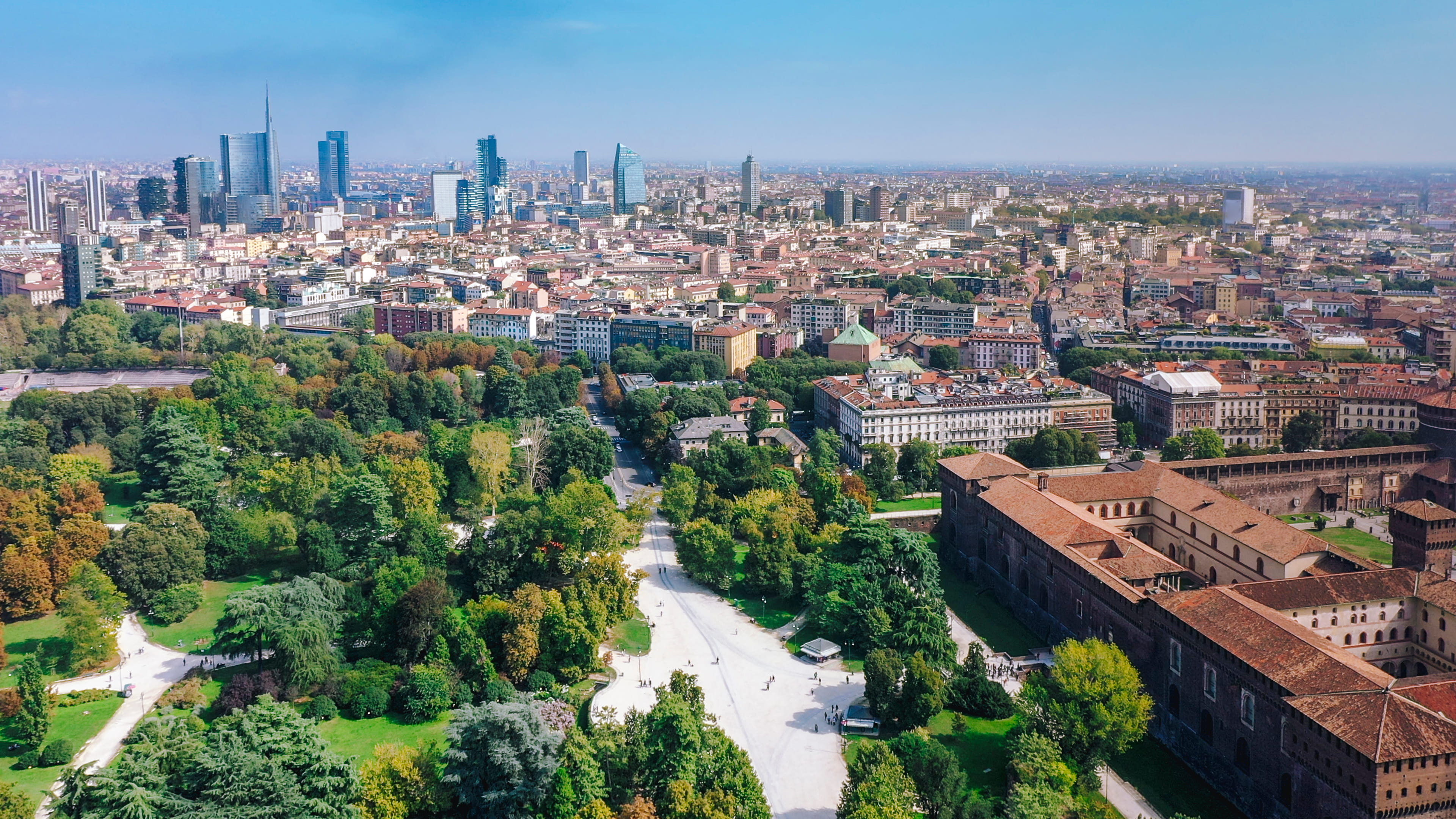 Aerial view of a large green park with paths and trees in the foreground and modern skyscrapers with city buildings in the background under a clear blue sky.