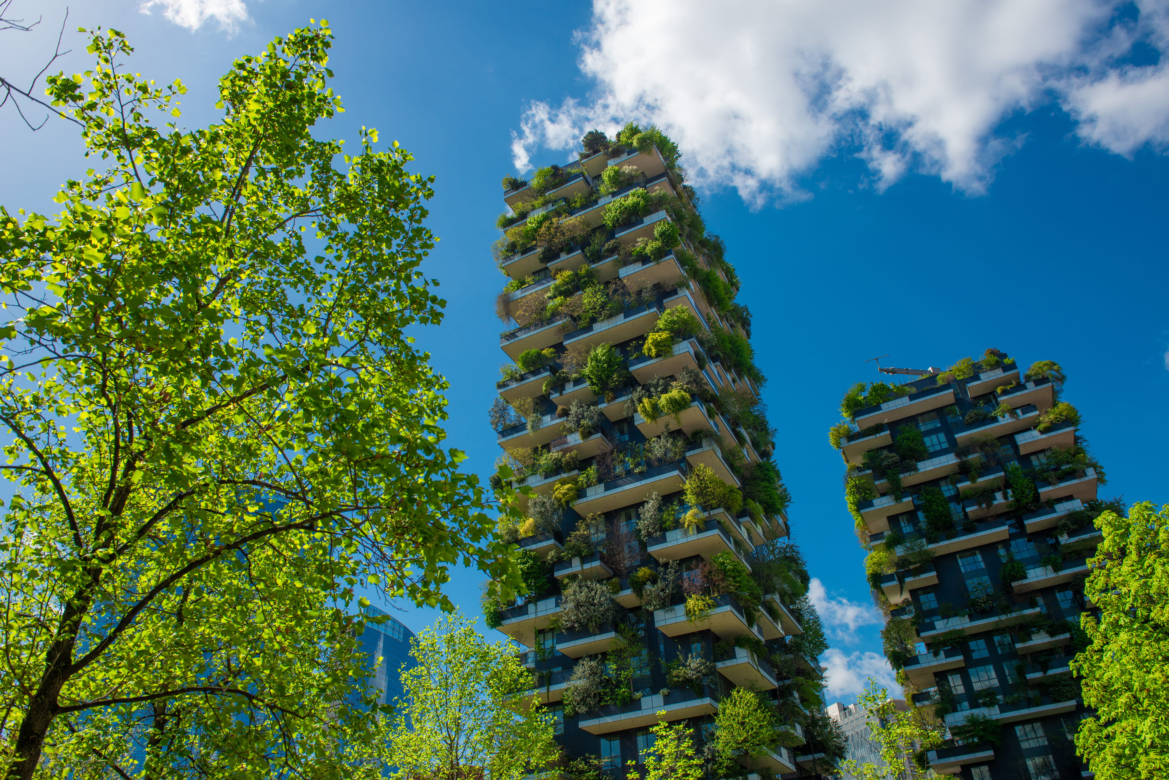 Two tall modern buildings covered with balconies filled with green plants under a blue sky with clouds.