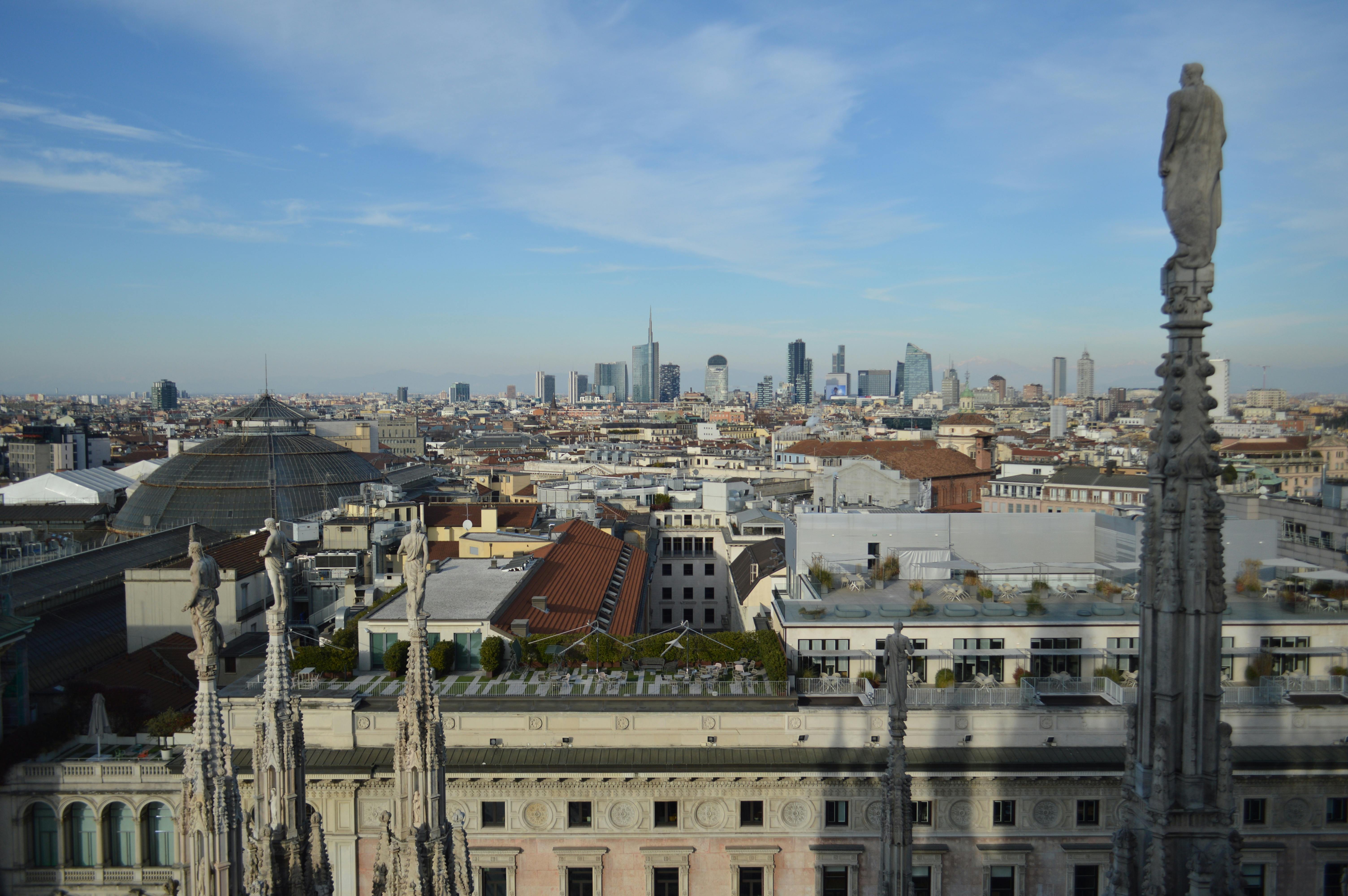 View over Milan cityscape with historic cathedral spires in foreground and modern skyscrapers in the distance under a partly cloudy sky.