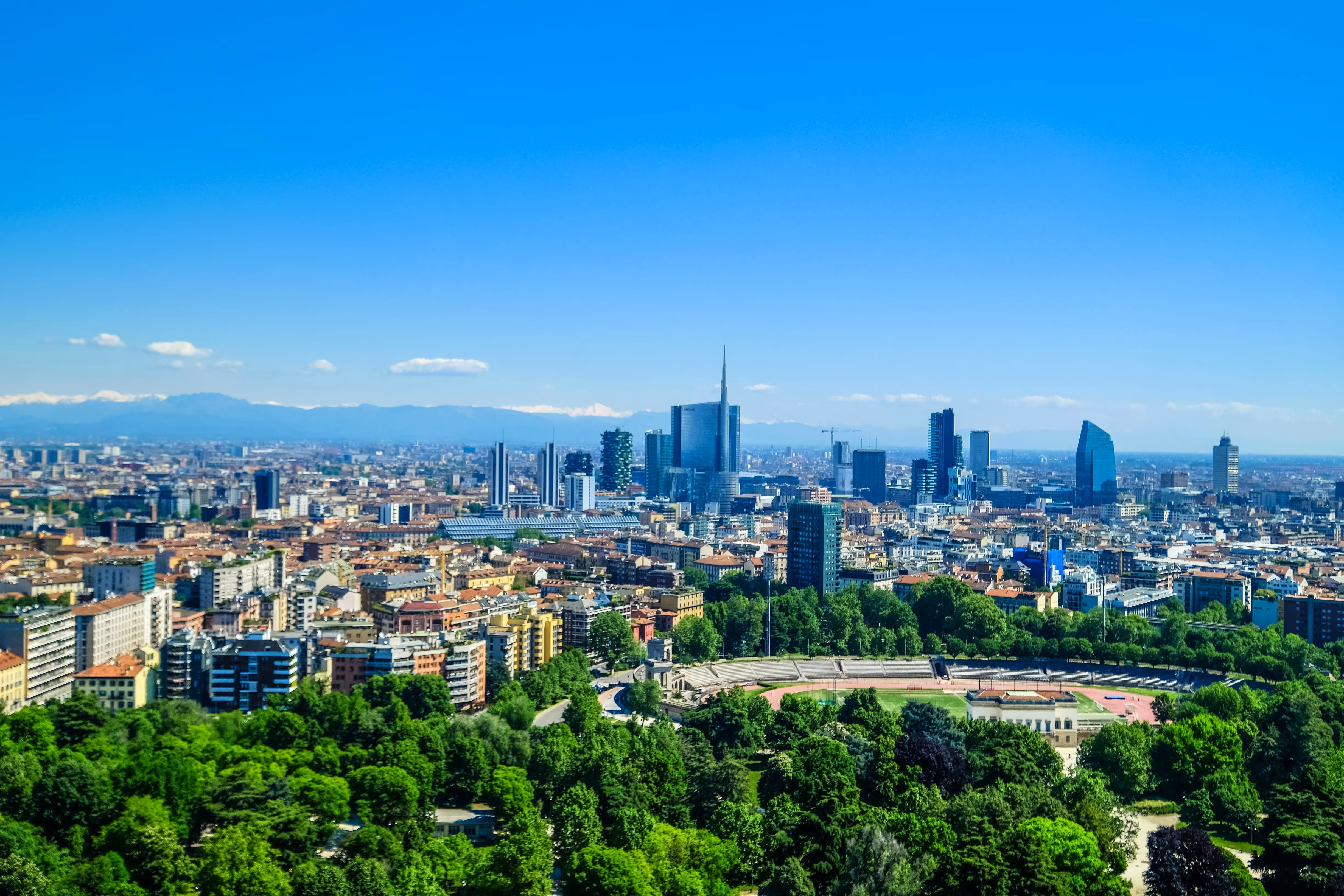 Panoramic view of Milan city skyline with modern skyscrapers and a lush green park under a clear blue sky.