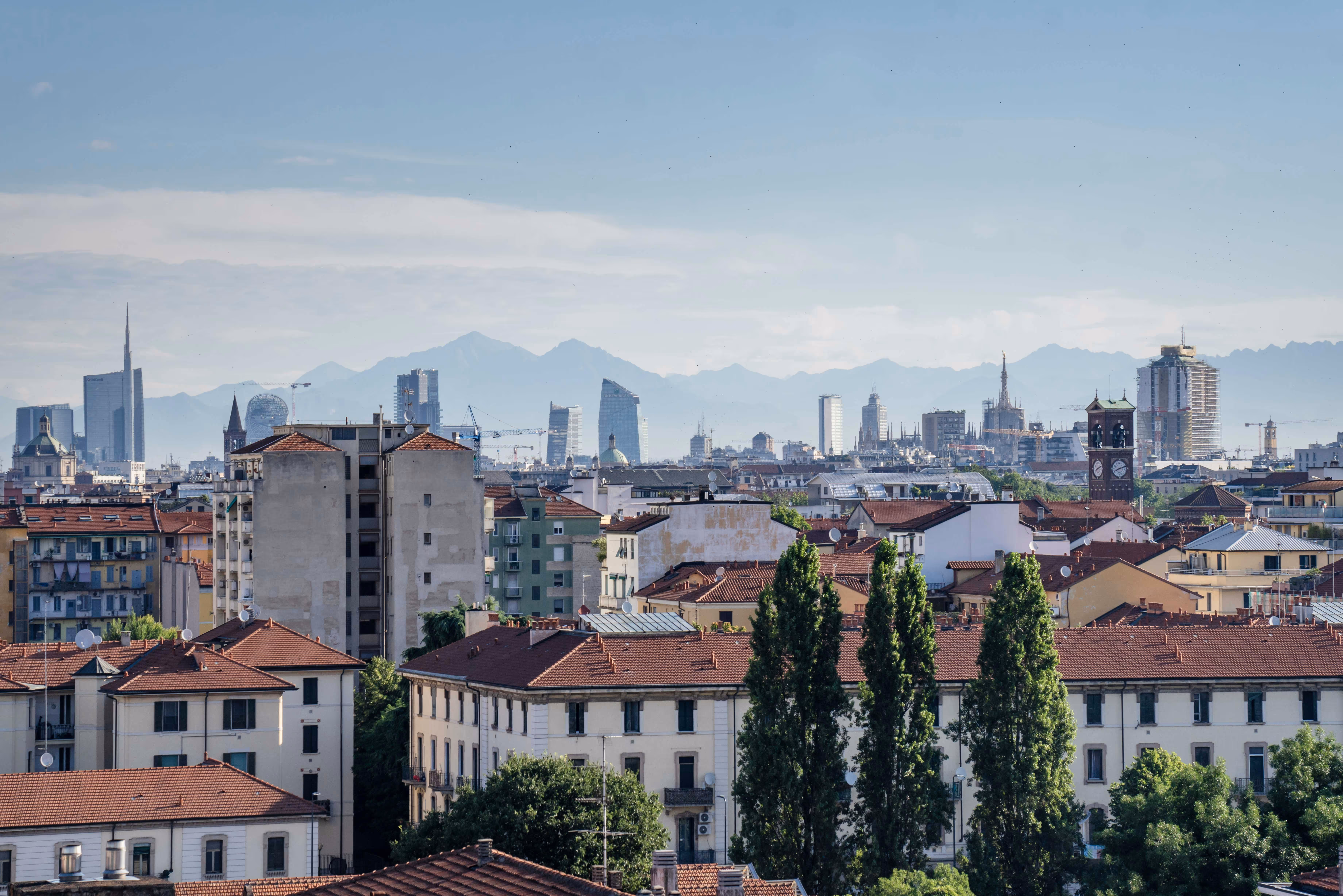 Panoramic view of Milano cityscape with red-roofed buildings, modern skyscrapers, and mountains in the background under a clear sky.