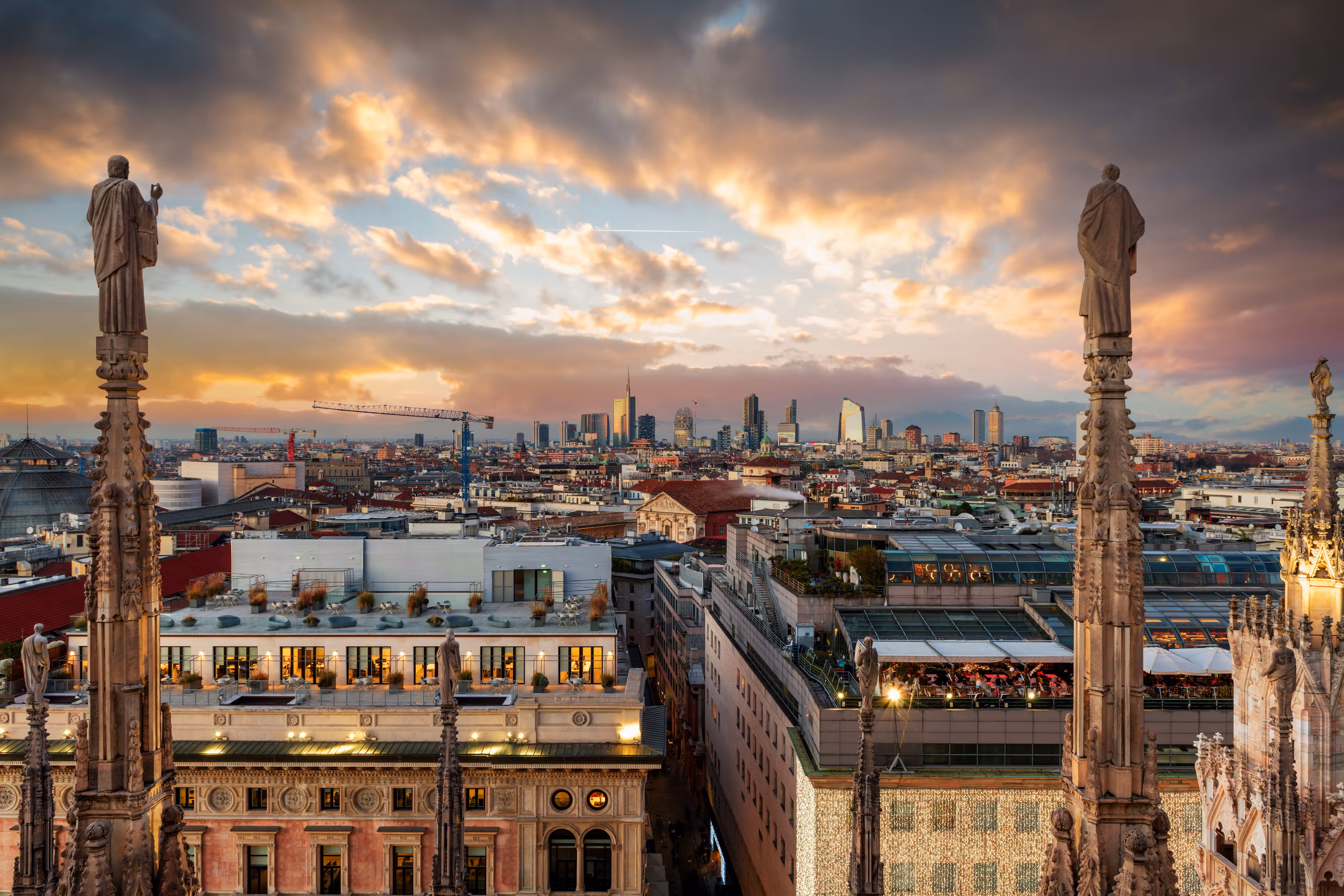 Sunset view of Milan skyline with statues atop gothic cathedral spires in the foreground and modern buildings in the background.