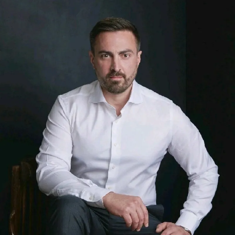 Homme sérieux en chemise blanche assis sur une chaise devant un fond sombre.