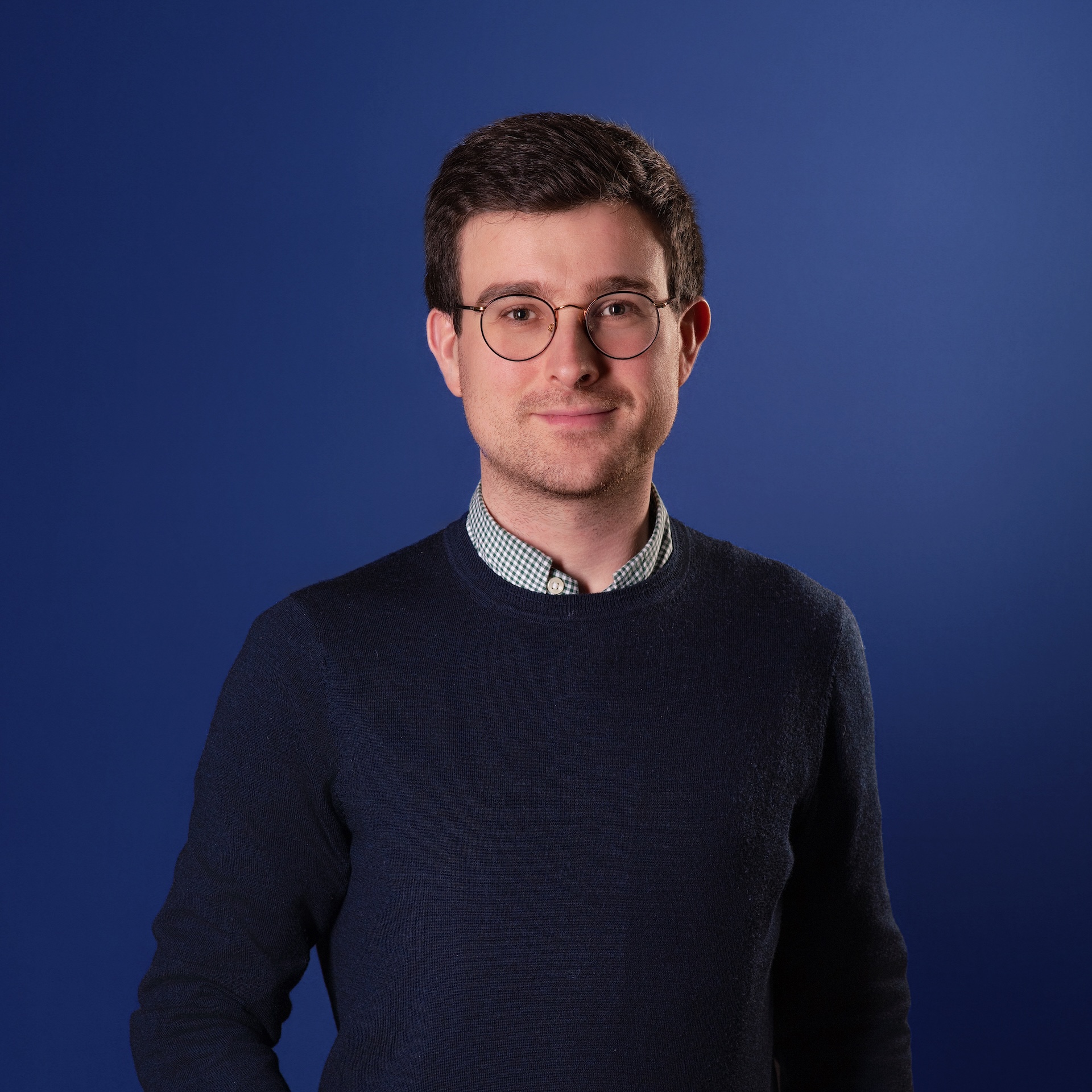 Homme portant un pull bleu marine et une chemise à carreaux avec des lunettes rondes sur fond bleu.