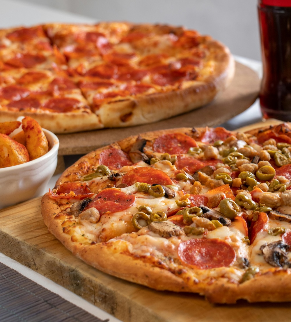 Two pizzas on wooden boards, one topped with pepperoni, mushrooms, green olives, and sausage, and a side bowl of seasoned potato wedges with a dark drink in the background.