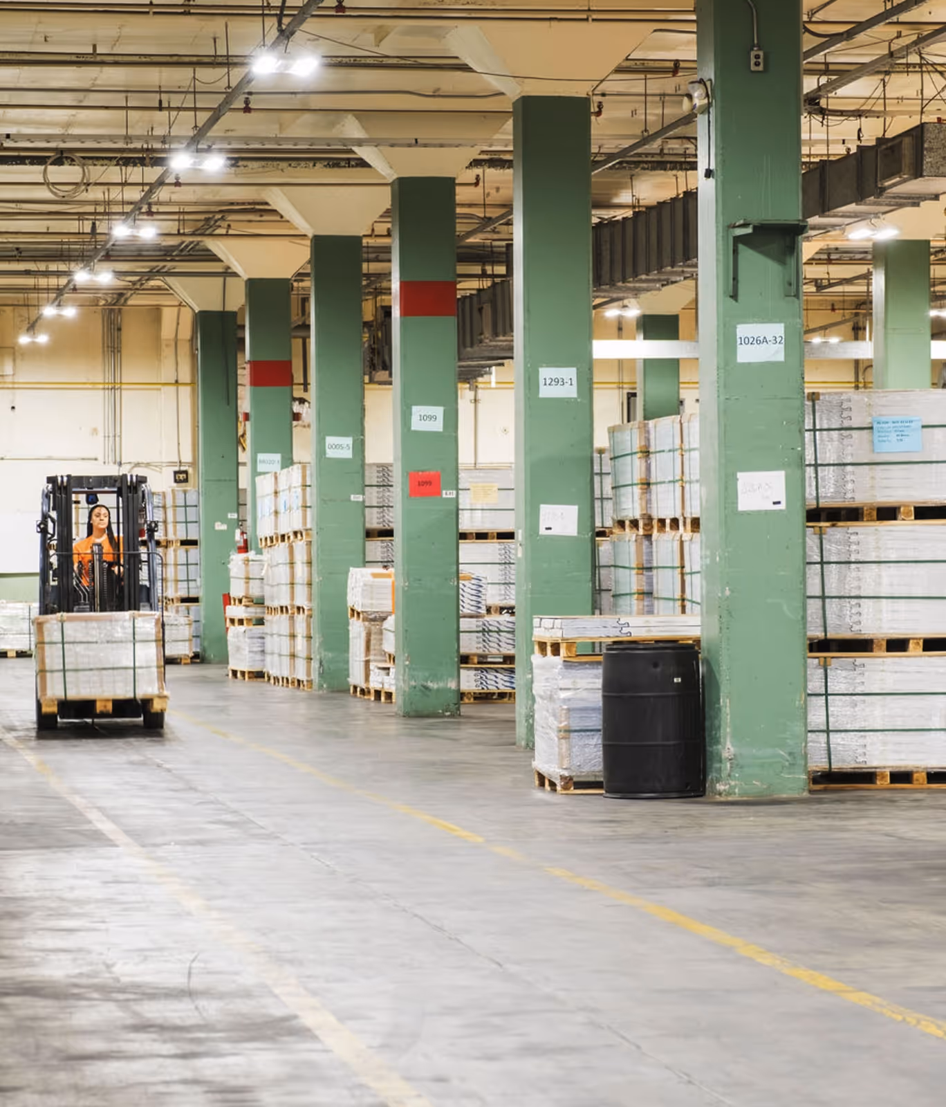 Warehouse interior with green columns, stacked pallets of goods, and a person operating a forklift.