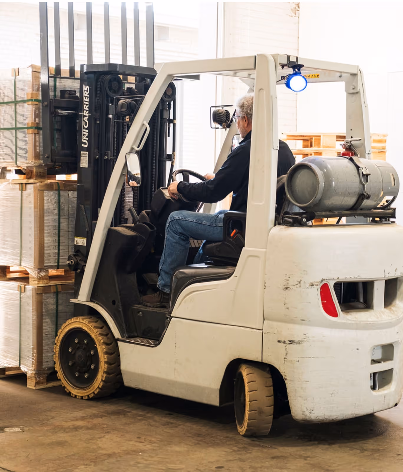 Worker driving a white forklift lifting stacked pallets wrapped in plastic inside a warehouse.