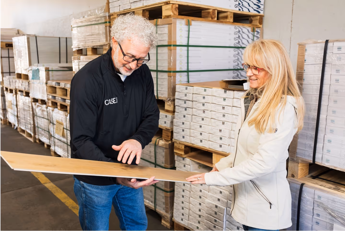 Man and woman examining a wooden plank sample in a warehouse filled with stacked boxes of flooring materials.