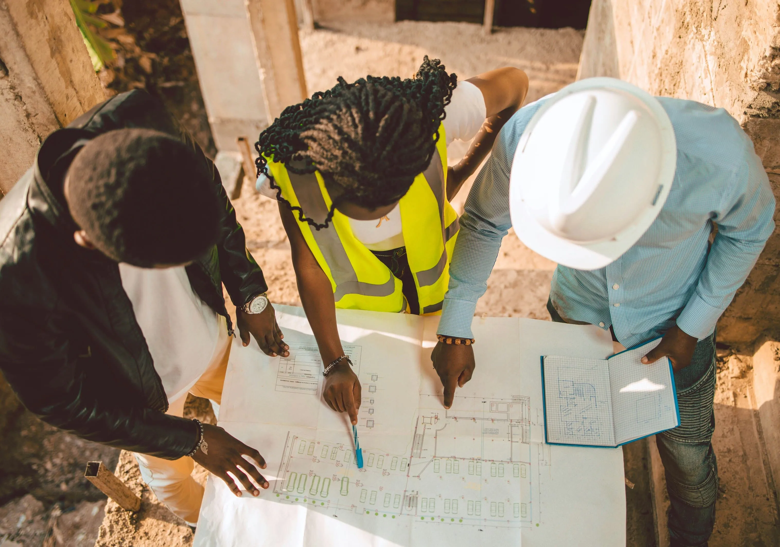 Trois professionnels de la construction examinent un plan et un carnet de notes sur un chantier ensoleillé.