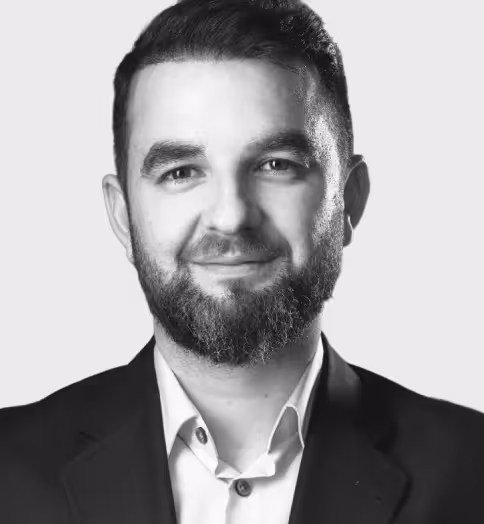 Smiling man with short dark hair and a full beard wearing a dark suit jacket and white shirt with an open collar on a light gray background.