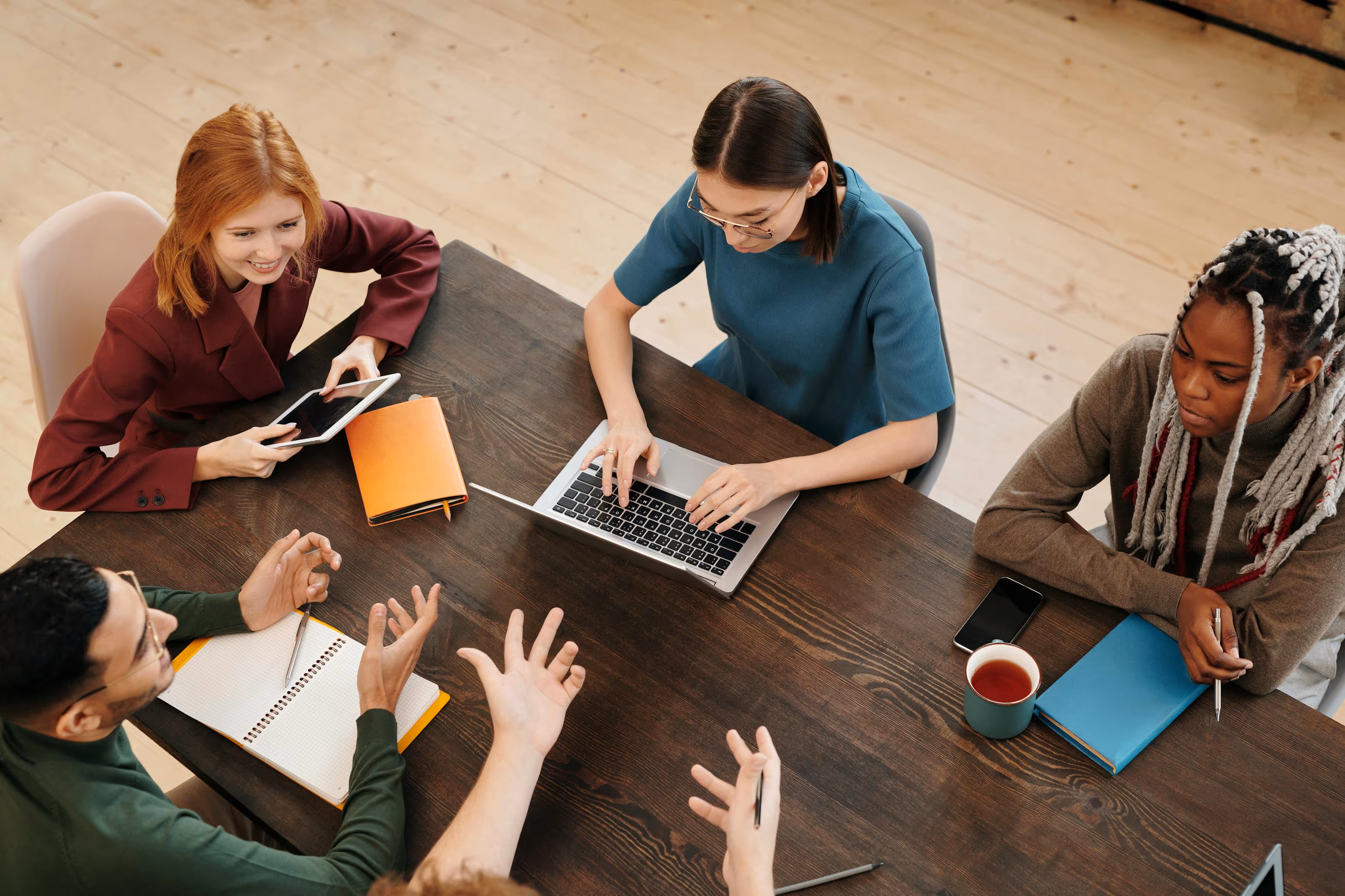 Five diverse people are collaborating at a wooden table with laptops, notebooks, and a cup. They appear engaged and focused, fostering a productive atmosphere.