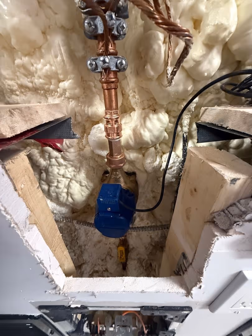 Foam-insulated wall cavity with copper plumbing and a blue water meter attached to copper pipes.