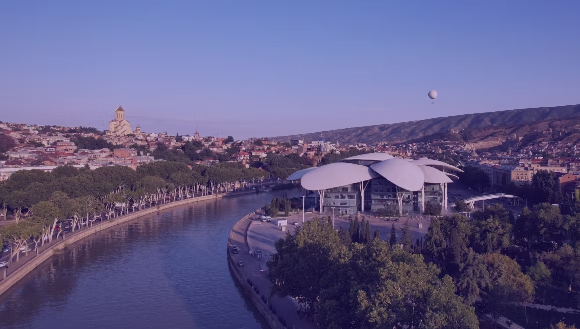 Modern building with petal-like roof by a river in a city. Trees line the riverbank. A balloon floats in the clear blue sky.