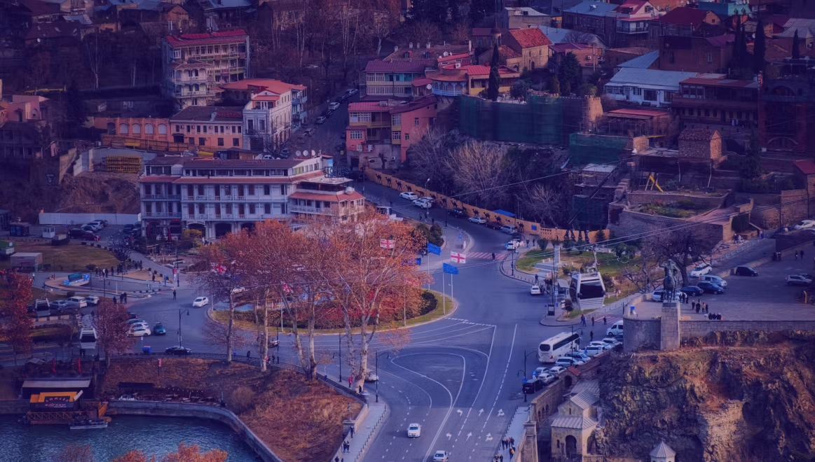 Aerial view of a city intersection with cars and pedestrians. Colorful buildings, trees, and a river in the background create a lively scene.