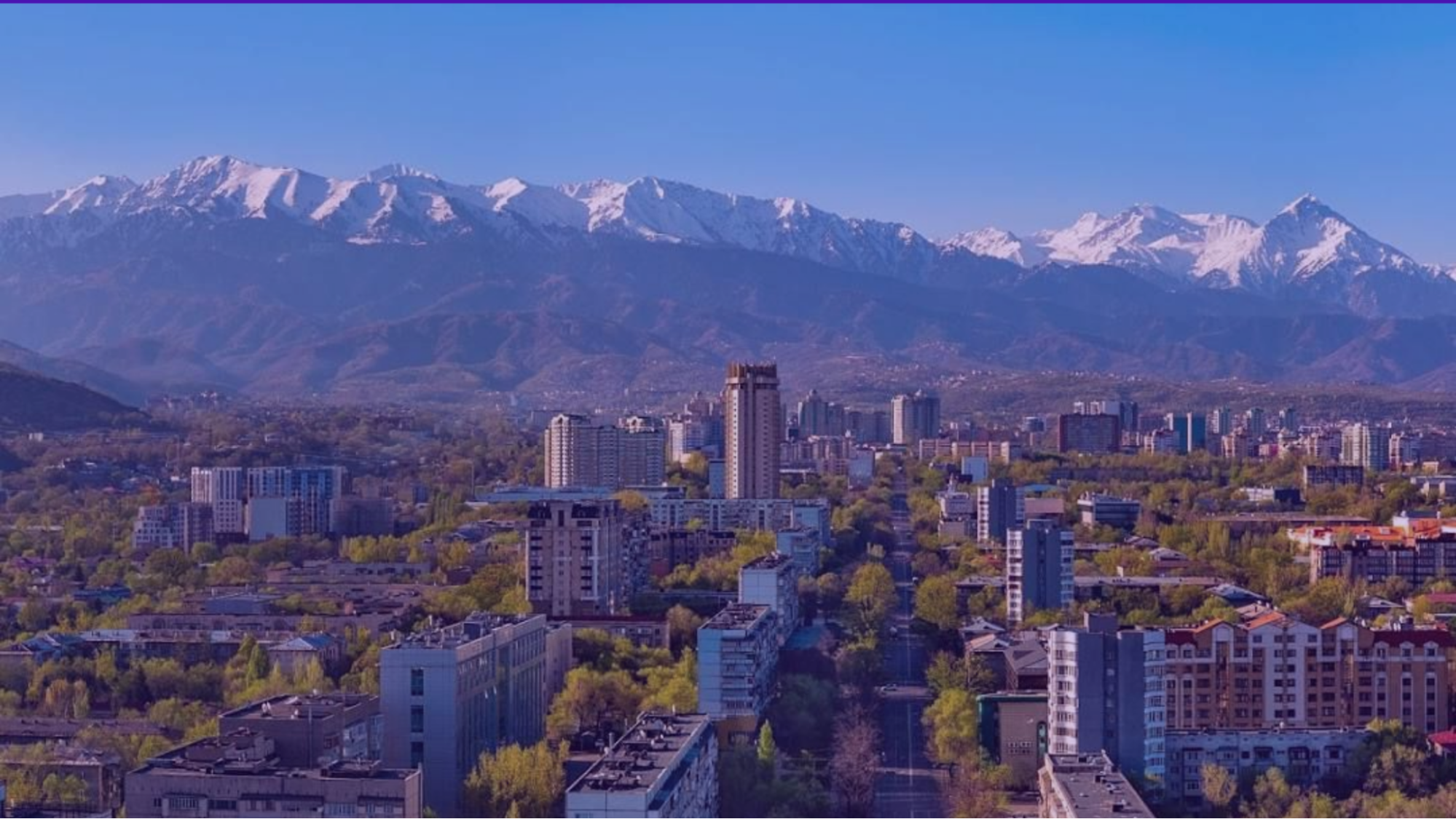 Cityscape with tall buildings and lush greenery under snow-capped mountains. Clear blue sky, setting an expansive and serene mood.