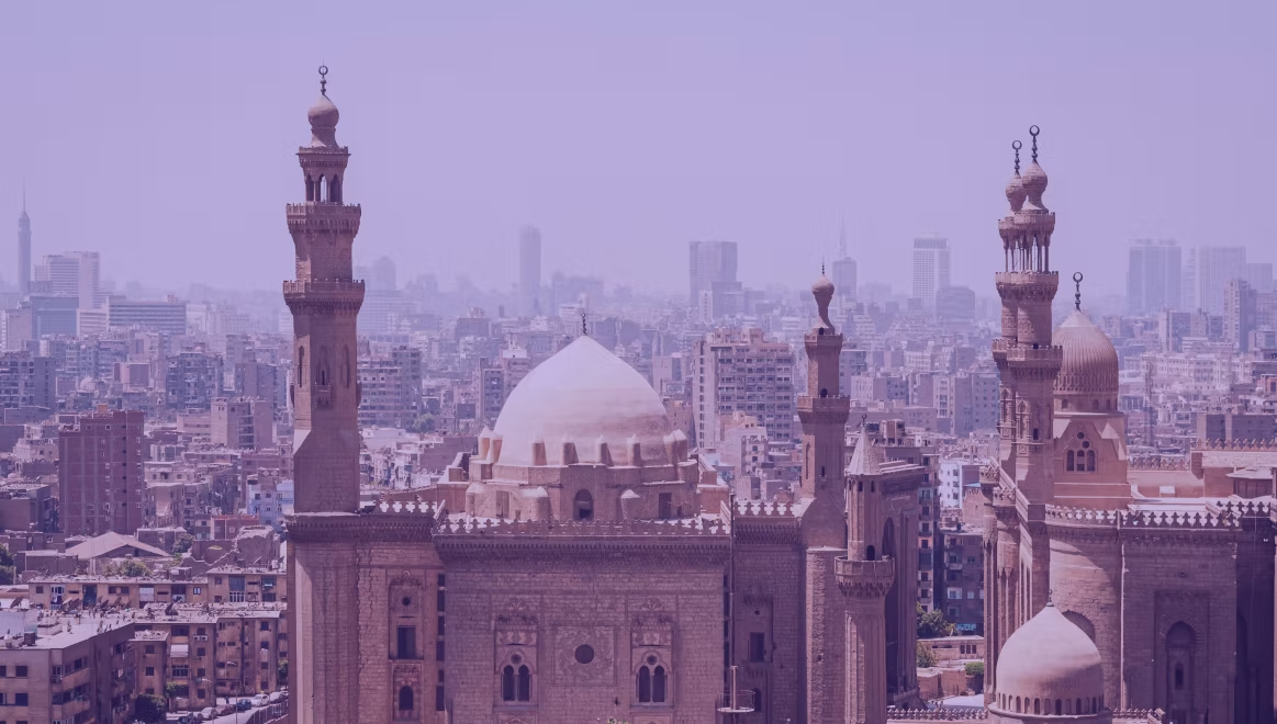 Skyline of Cairo featuring the historic Sultan Hasan Mosque and its minarets, with modern buildings in the hazy background, under a purple hue.