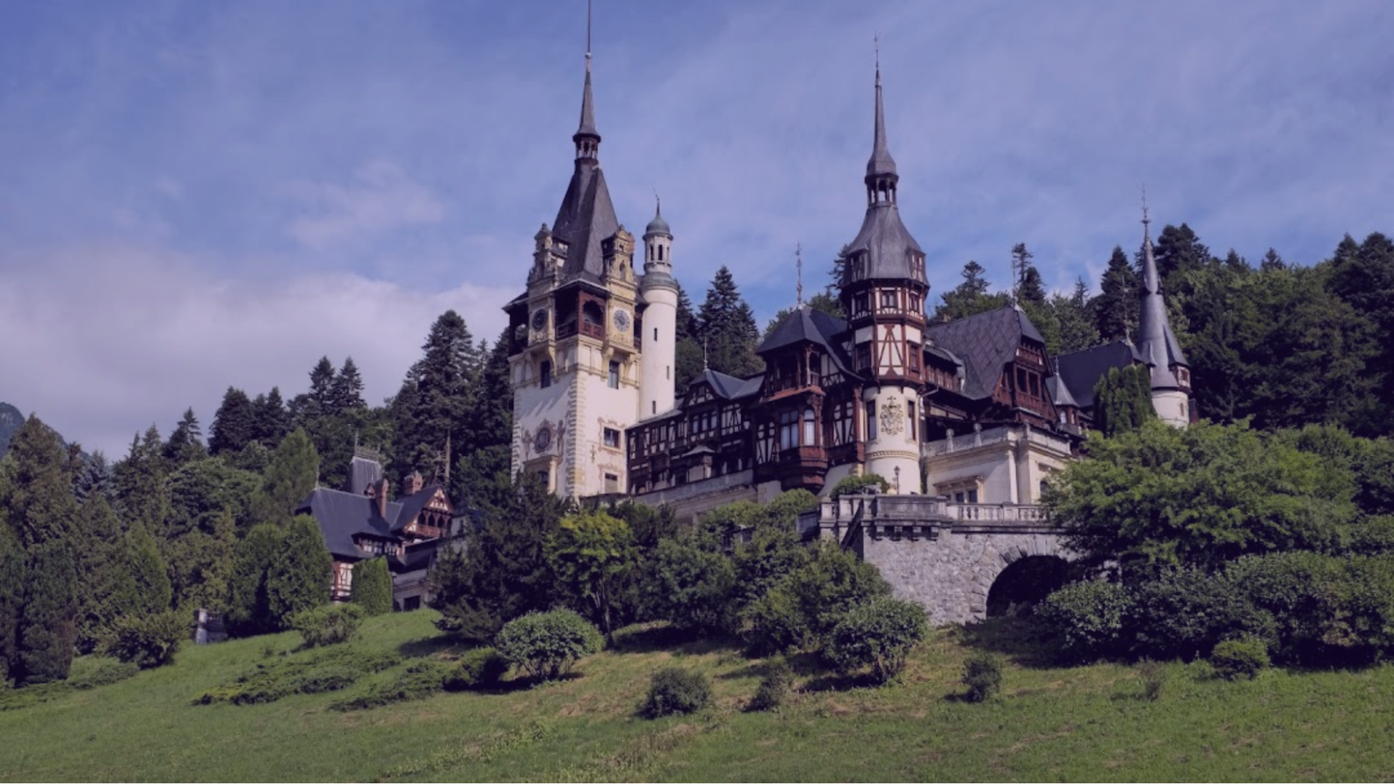 A grand castle with multiple spires on a lush green hillside, surrounded by dense trees. A bright, clear sky enhances the majestic view.
