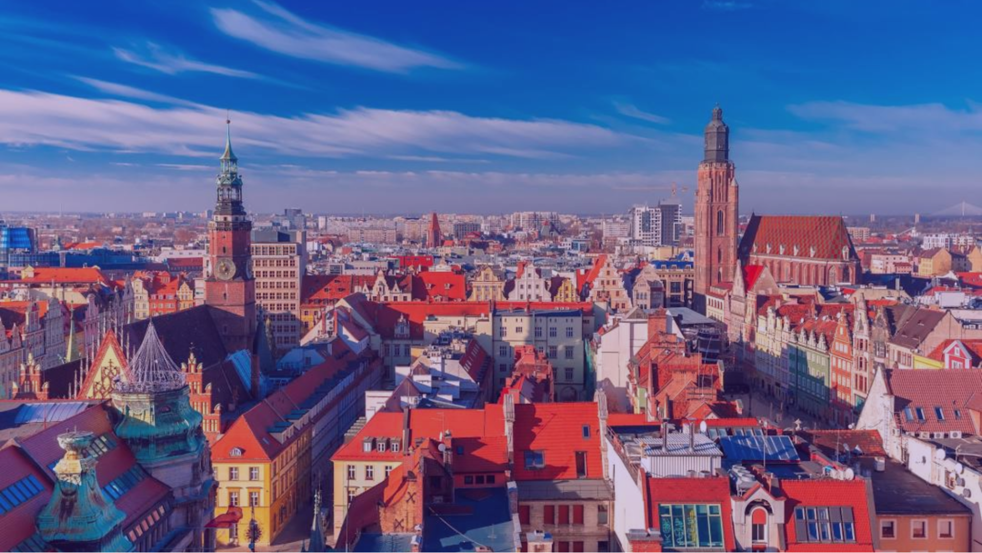 Aerial view of a cityscape with colorful buildings and two tall towers under a vivid blue sky with streaked clouds.