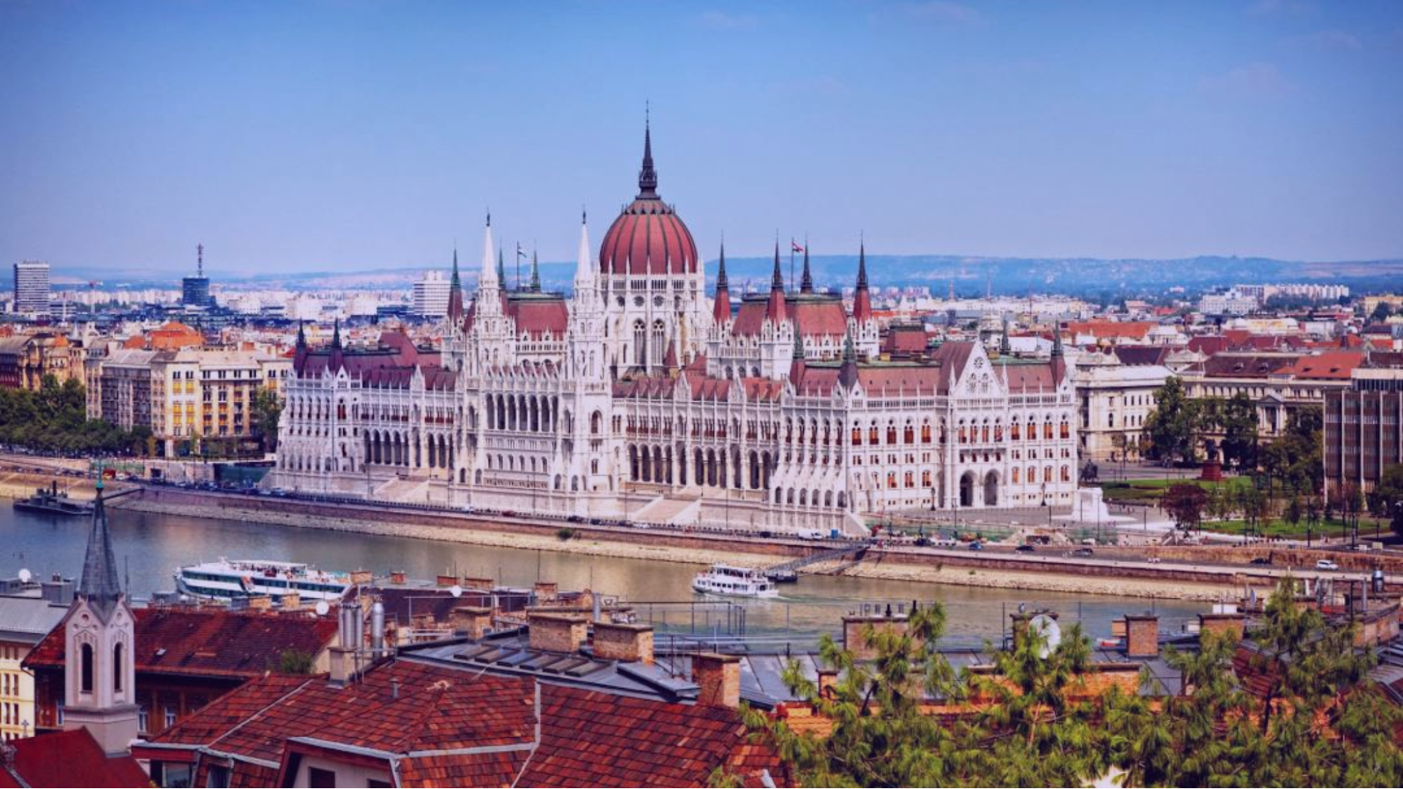 The Hungarian Parliament Building with red domes and spires overlooks the Danube River. Boats traverse the water under a clear blue sky.