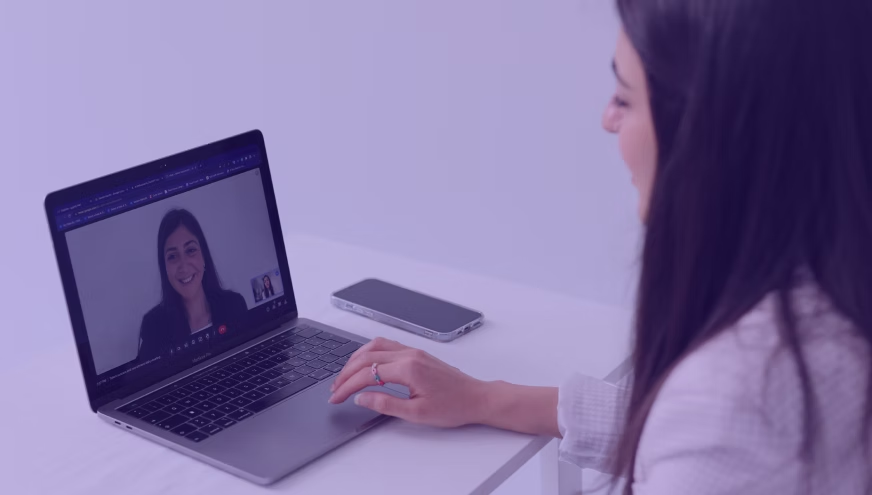 Woman on video call via laptop, smiling. Smartphone on white desk. Room has a purple tint. Mood is professional and friendly.