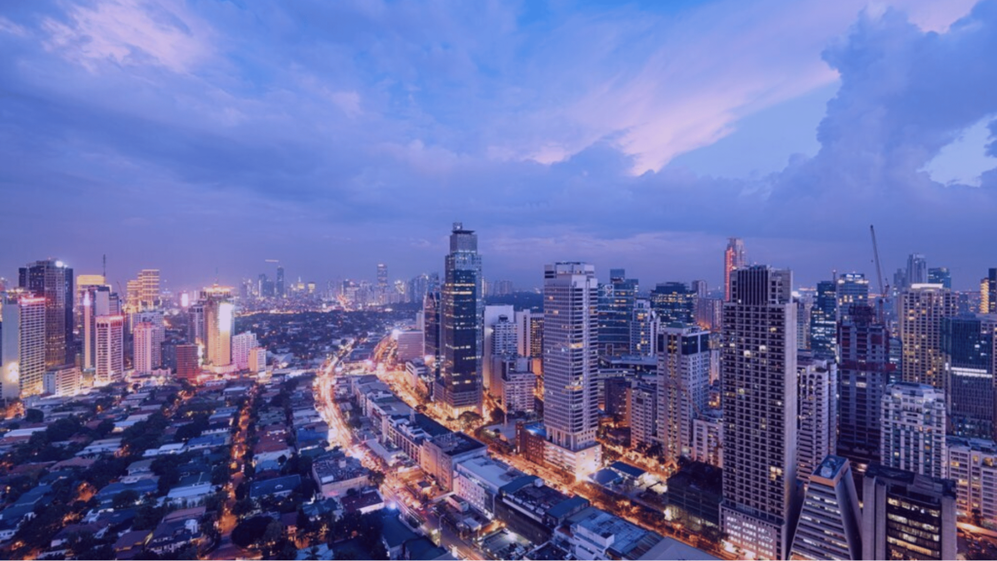 Twilight city skyline with glowing skyscrapers and winding traffic below under a blue-purple cloudy sky.