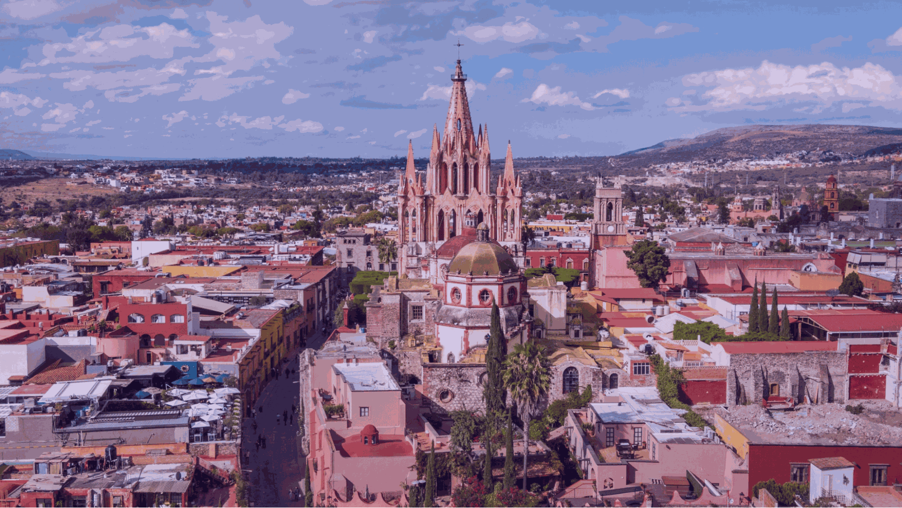 Aerial view of a colorful city with a pink Gothic cathedral spire rising above rooftops under a cloudy blue sky.