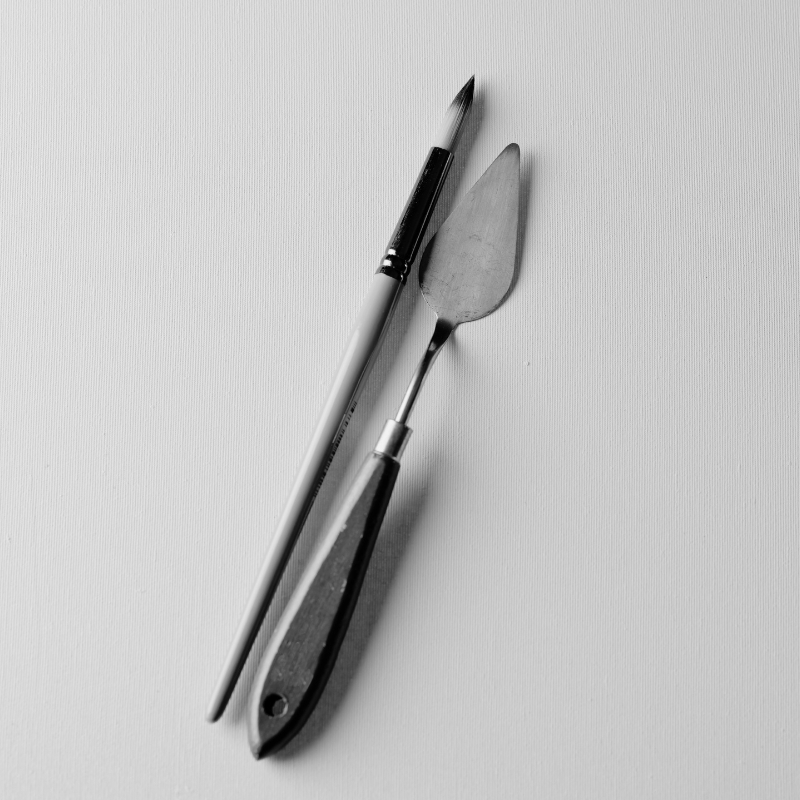 A paintbrush and a palette knife placed side by side on a blank canvas.