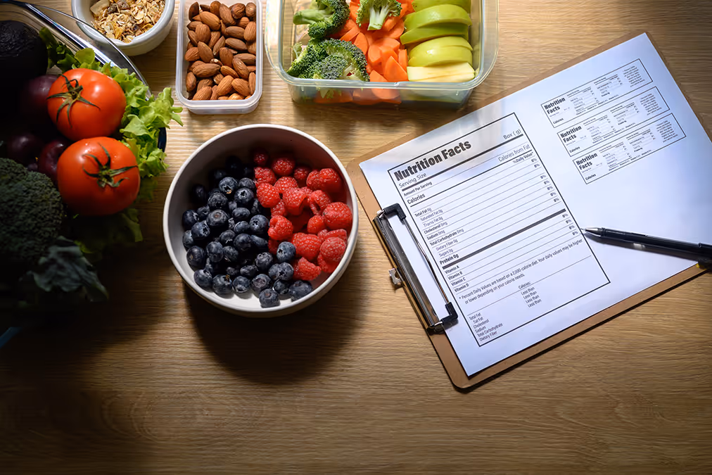 Assorted fresh fruits, vegetables, nuts, and nutrition facts on a clipboard with pen on wooden table.