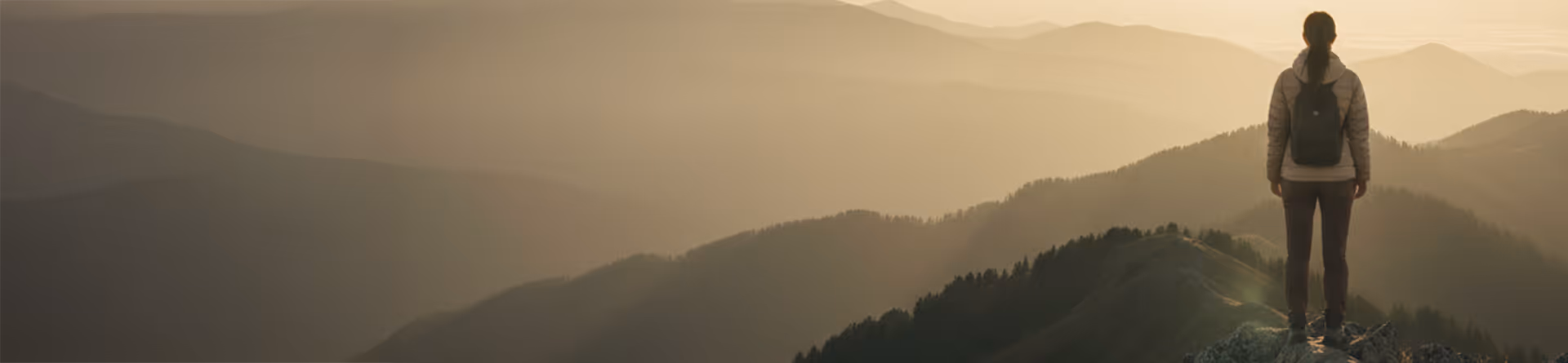 Person with a backpack stands on a rock overlooking a misty mountain range at sunrise.