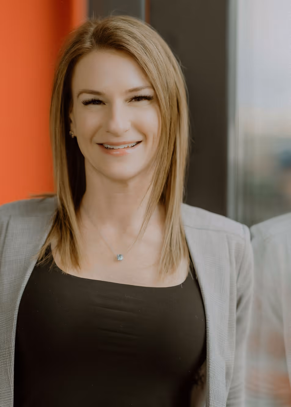 Smiling woman with straight blonde hair wearing a gray blazer and black top, standing indoors near a window.