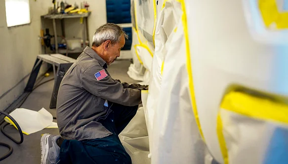 Painter in a spray suit at the paint booth preparing a bumper for refinishing