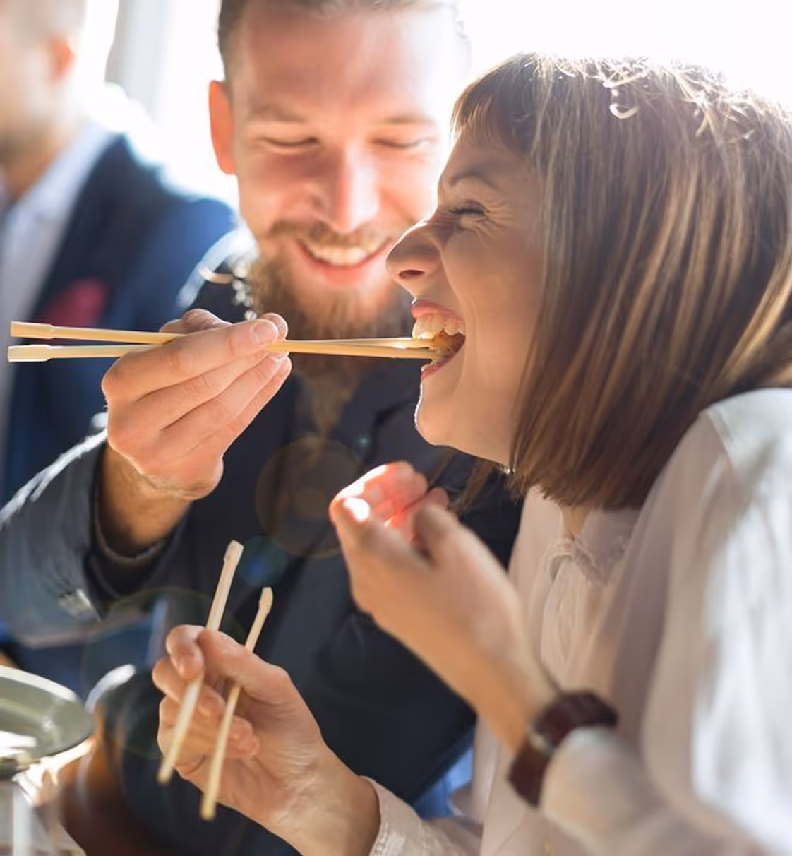Man feeding woman a bite of food with chopsticks while both smile.