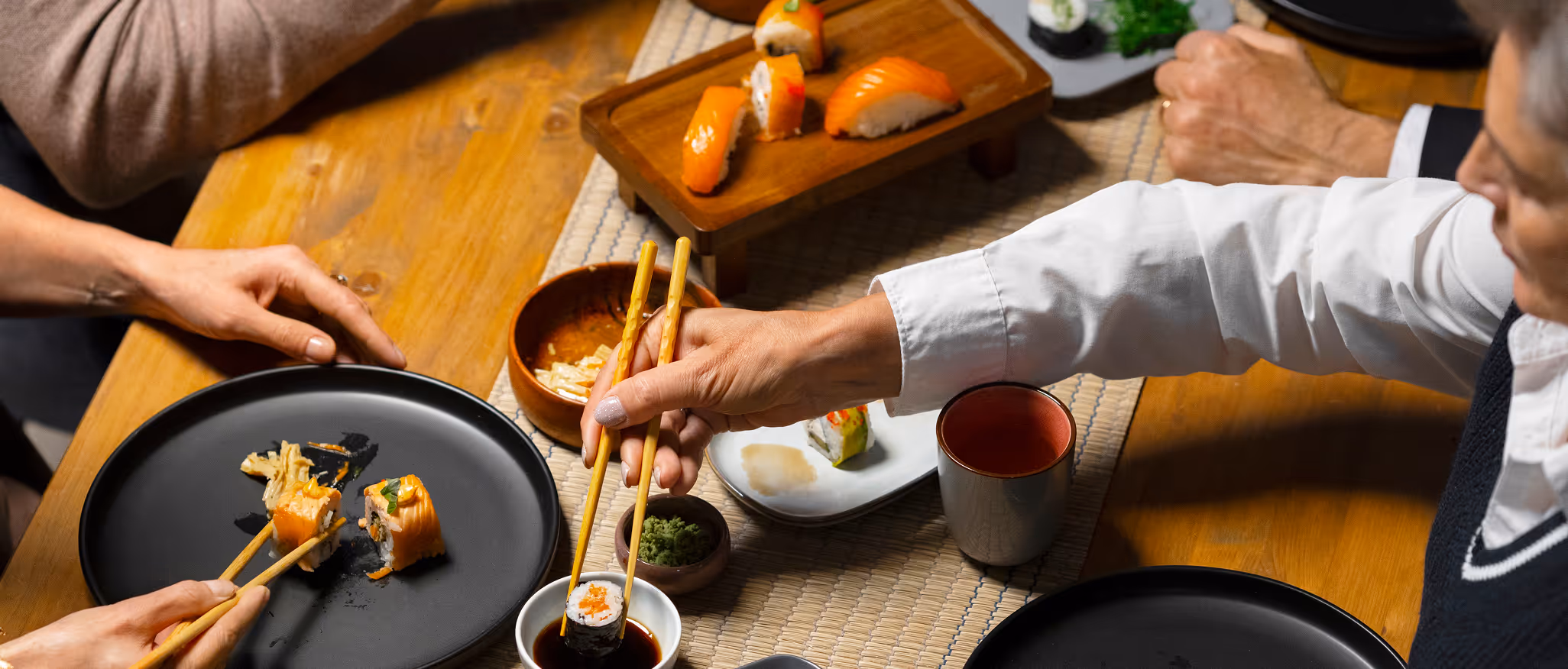 Two people using chopsticks to eat sushi at a wooden table with soy sauce and wasabi.