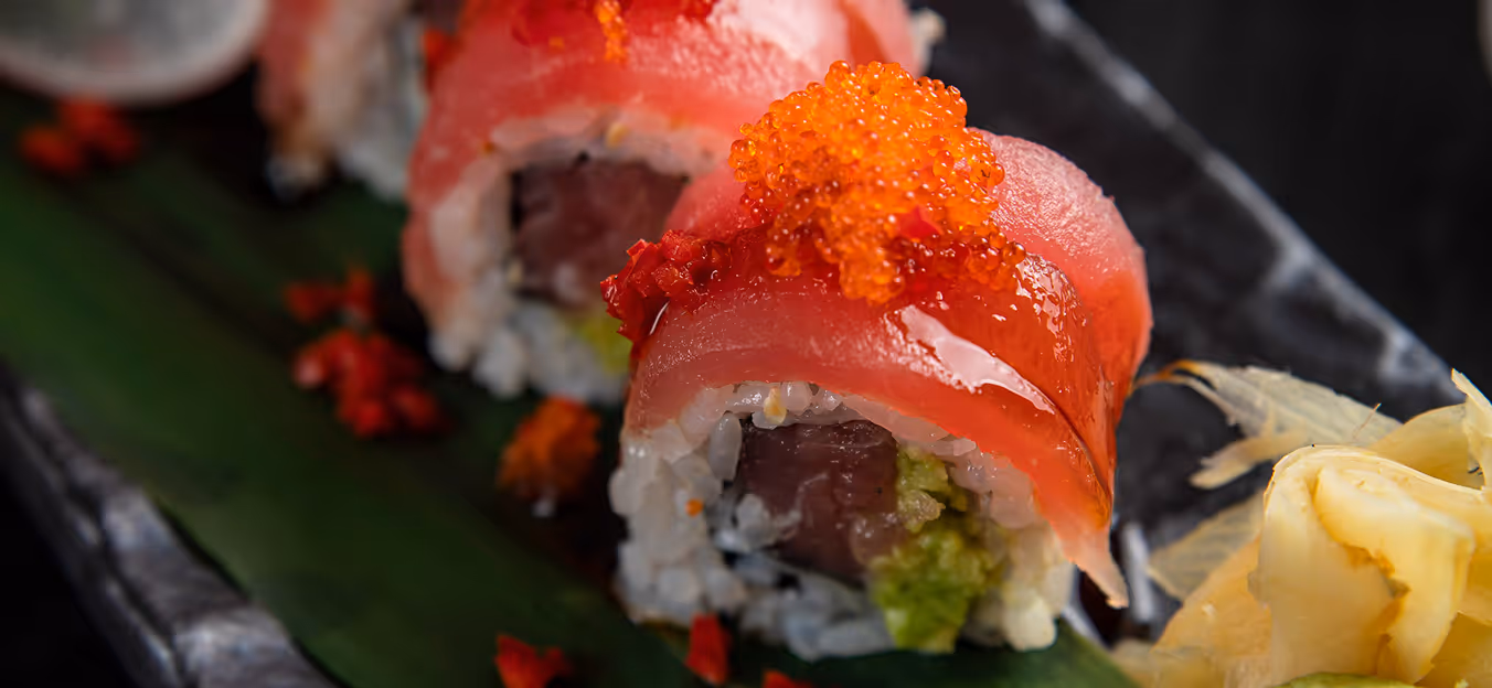 Close-up of sushi rolls topped with red fish, orange fish roe, and served with ginger on a dark plate with a green leaf.