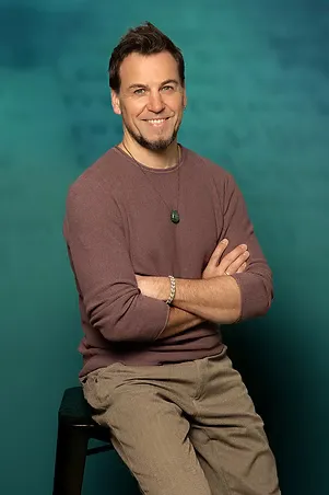 Smiling man with short dark hair and a goatee, wearing a brown sweater and beige pants, sitting on a black stool with arms crossed.