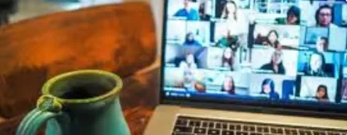 Blue ceramic mug on a wooden table next to an open laptop showing a virtual meeting with multiple participants.