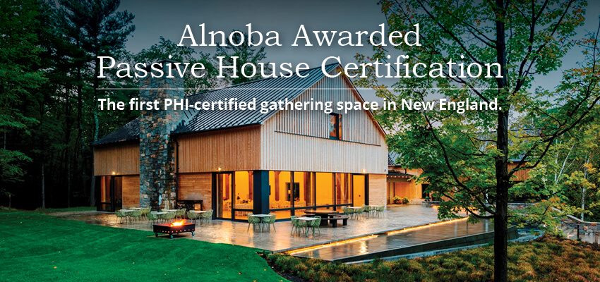 Exterior of Alnoba's award-winning Passive House gathering space in New England, showing wood siding, stone features, lit interiors, outdoor furniture, fire pit, and landscaped grounds. 