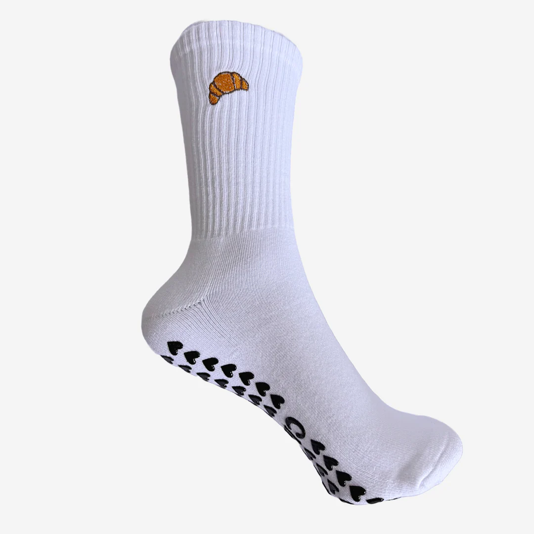 White sock with a small orange croissant embroidery near the top and black heart-shaped grips on the sole.