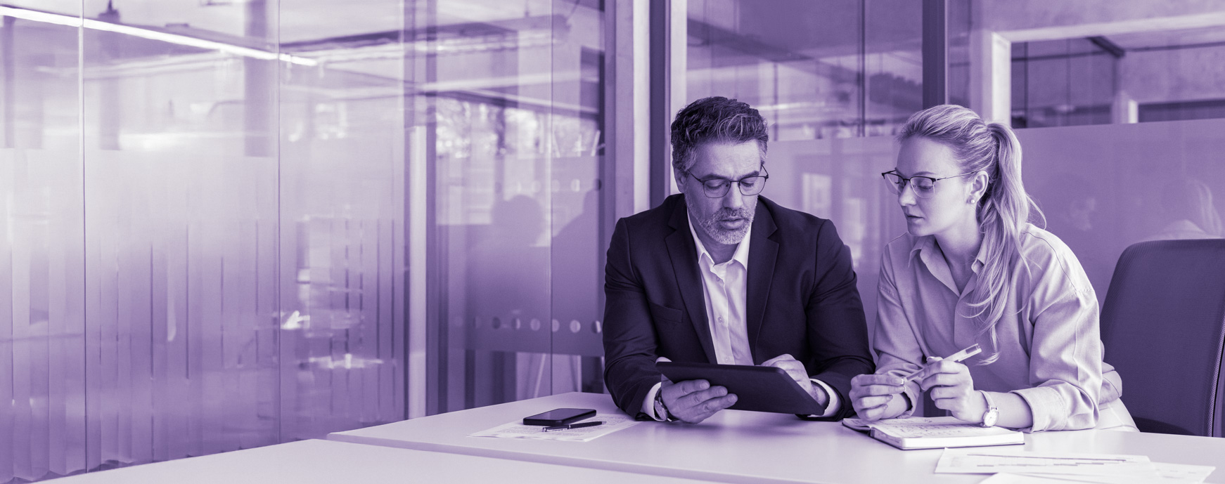 Two business professionals wearing glasses reviewing a tablet together in a modern office conference room.
