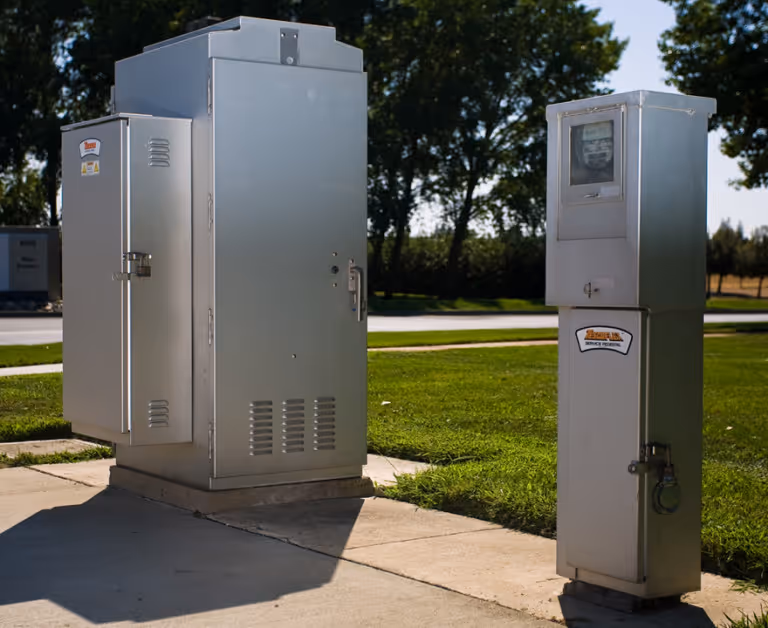 Two silver outdoor electrical utility cabinets on a concrete surface with grass and trees in the background.