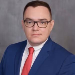 Man wearing glasses, a blue suit, white shirt, and red tie, posing in front of a plain gray background.