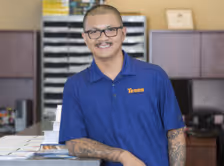 Smiling man with glasses and tattoos wearing a blue polo shirt leaning on a desk in an office.