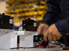 Hands assembling or adjusting black electronic components on a white workbench in a manufacturing environment.