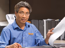 Man in blue shirt wearing glasses, holding and reviewing large papers in an office setting with printers in the background.