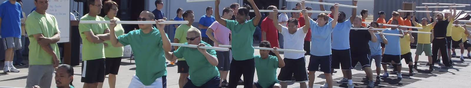 Group of people outdoors performing squats while holding long poles at shoulder height, dressed in colorful t-shirts.
