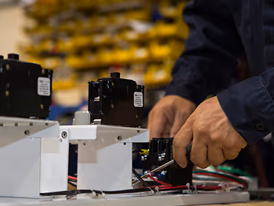 Hands of a person assembling or wiring electrical components on a white panel in an industrial setting.