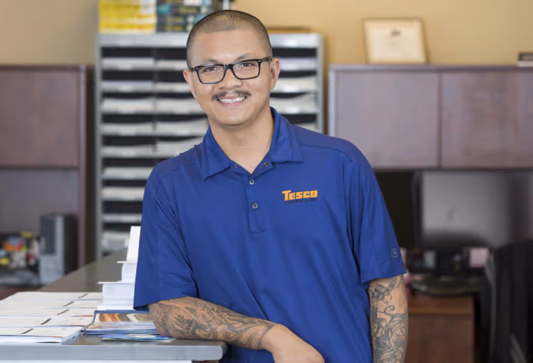Smiling man with glasses and tattoos leaning on an office counter wearing a blue Tesco Control Co polo shirt.