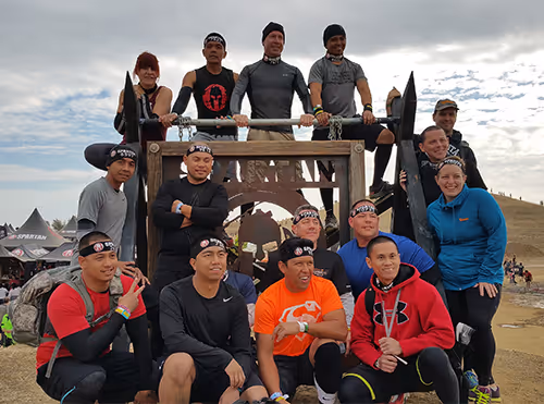 Group of people posing together at a Spartan Race event in front of a wooden Spartan sign outdoors under a cloudy sky.